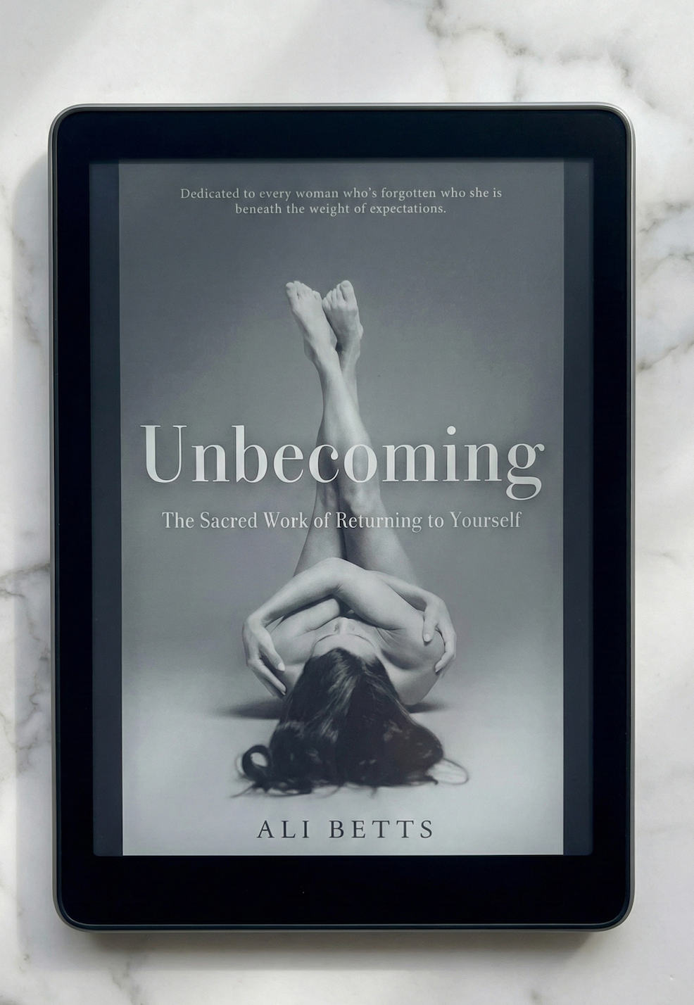 Ebook cover of 'Unbecoming' by Ali Betts on a tablet with a marble background